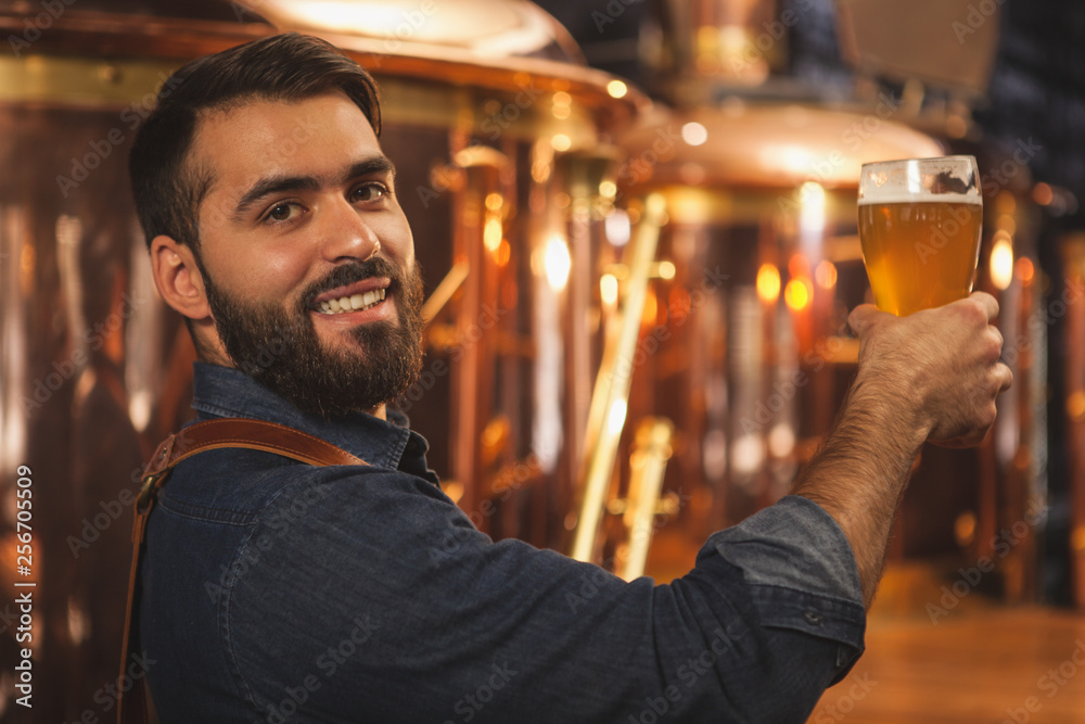 Happy bearded professional brewer smiling to the camera over his shoulder, holding glass of delicious beer, copy space. Attractive male bartender posing with a glass full of tasty beer. Bar, pub conce