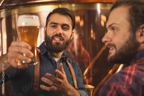 Obraz na płótnie Handsome bearded brewer smiling, talking to his colleague while examining freshly brewed craft beer