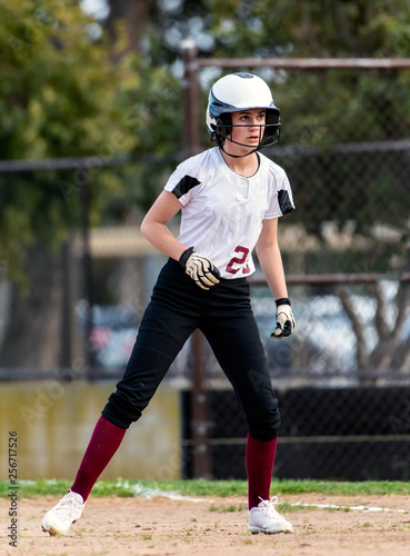 Female teen softball player in black and white uniform taking a cautious lead off from first base.