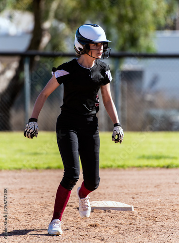 Female softball player in black uniform taking an agressive lead off second base and remaining alert during game. photo