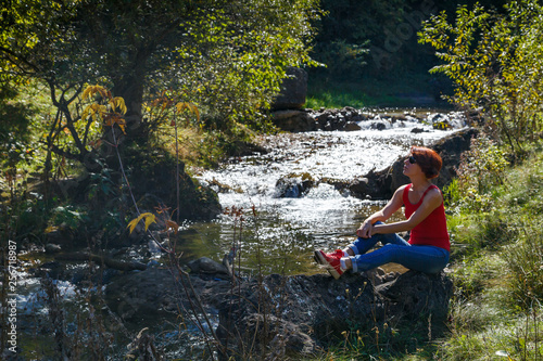 Fototapeta Naklejka Na Ścianę i Meble -  Young woman sits on a stone near a small stream on a warm autumn day
