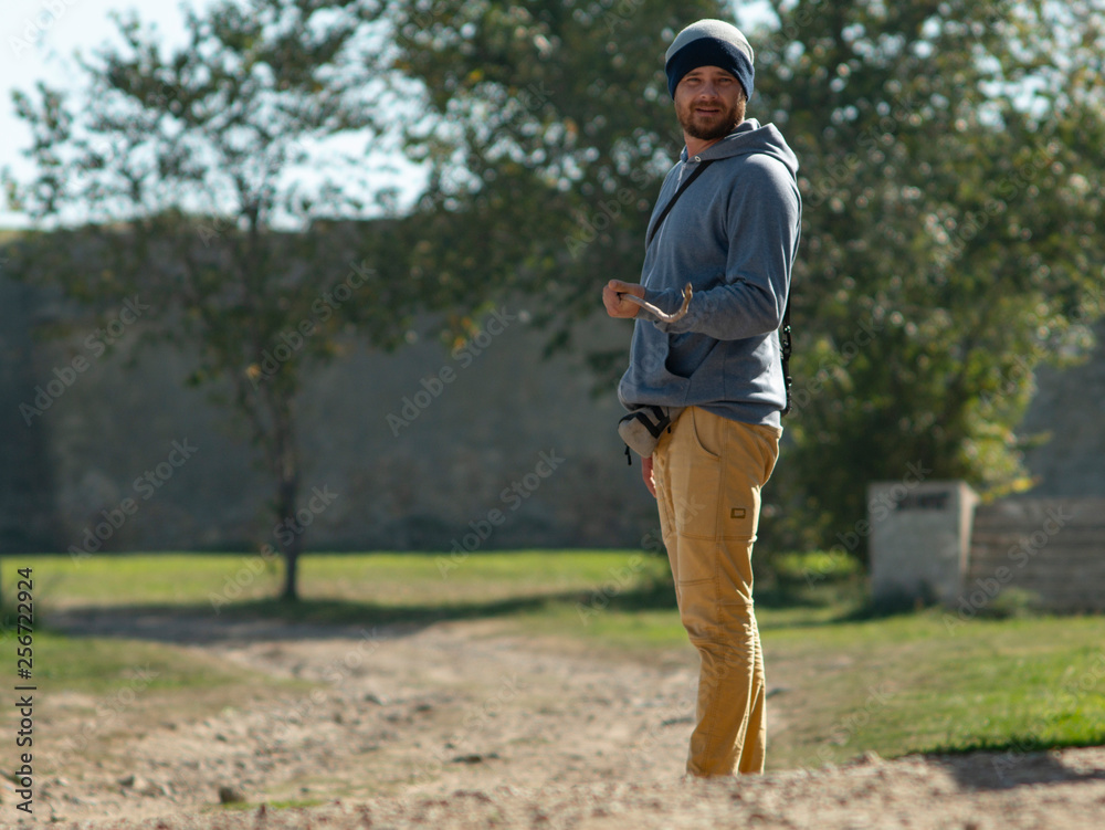 a man in a hooded sweater and knitted hat is standing against a background of green trees with a stick and smiling