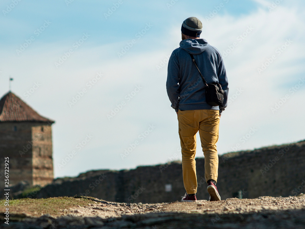 a man in a hooded sweater and knitted hat is walking along the road to the fortress against the background of the tower and the blue sky
