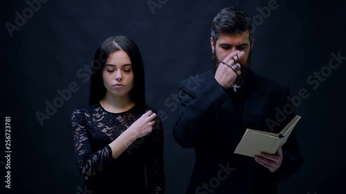 Girl and priest dressed in black baptized on a black background. Priest prays on a black background. Religious nun in religion concept against dark background. religion opium for the people.