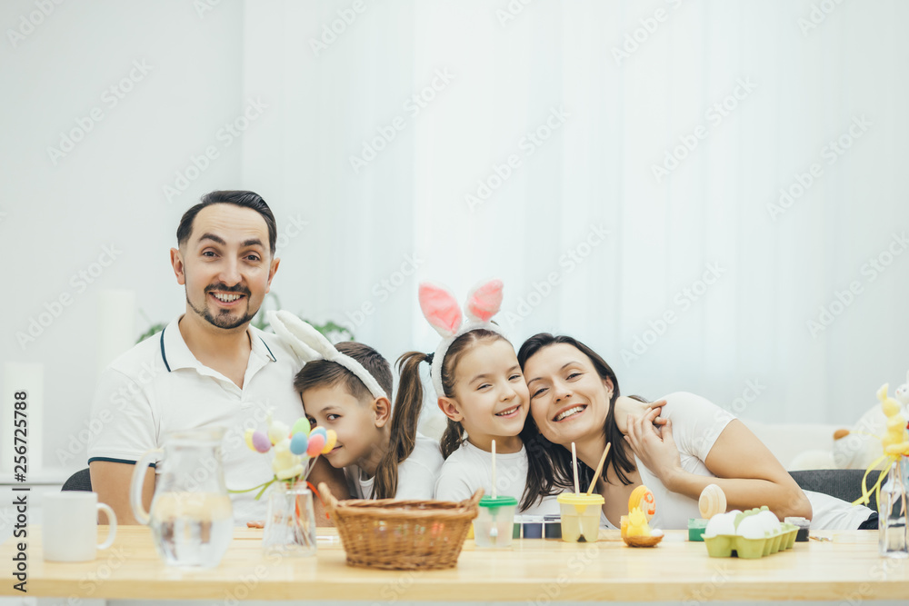 Happy smiling family preparing for Easter. Son hugs father, daughter hugs mother. Children in bunny ears. A lot of holiday tiny things on the table. Happy smiling family preparing for Easter. Son hugs father, daughter hugs mother. Children in bunny ears. A lot of holiday tiny things on the table.