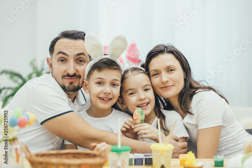 Happy family sitting at the table full og easter supplies  hugging and smiling. Children in funny bunny ears. Cute girl with pigtails is holding a colored egg.
