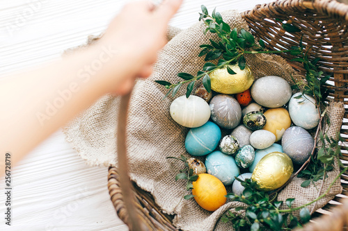 Hand holding  rustic wicker basket with stylish Easter eggs with green buxus branches on white wooden background. Easter hunt concept. Happy Easter.