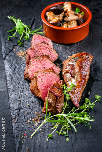 Traditional barbecue aged sliced venison sirloin with mushroom and herbs as closeup on a carbonized old board photo