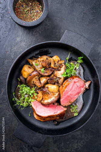 Traditional barbecue aged sliced venison backstrap with mushroom, onions and herbs as top view in a cast-iron pan