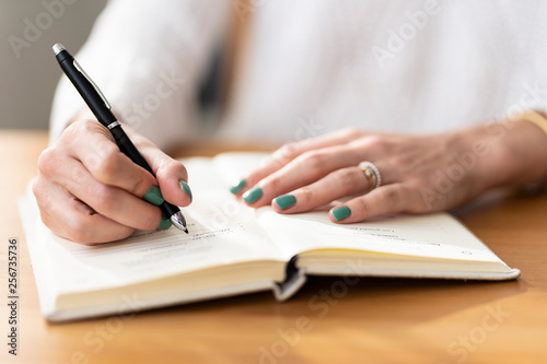 Primer plano de manos femeninas con u  as pintadas de verde  escribiendo con un bol  grafo negro elegante en un cuaderno o agenda sobre un escritorio de madera  en un entorno de oficina.