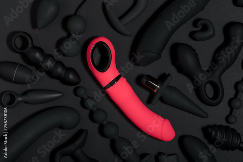 Erotic pleasure toys on a black background. Sex gadgets and masturbation devices. Pink dildo surrounded by black rubber and silicone sex toys: anal butt plugs, vibrators, vaginal balls. Top view.