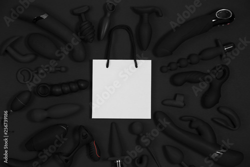 Shopping paper bag surrounded by black sex toys on a black background. Erotic pleasure toy. Sex gadget and masturbation device. Top view. Place for text. Flat lay. Sex shop concept.