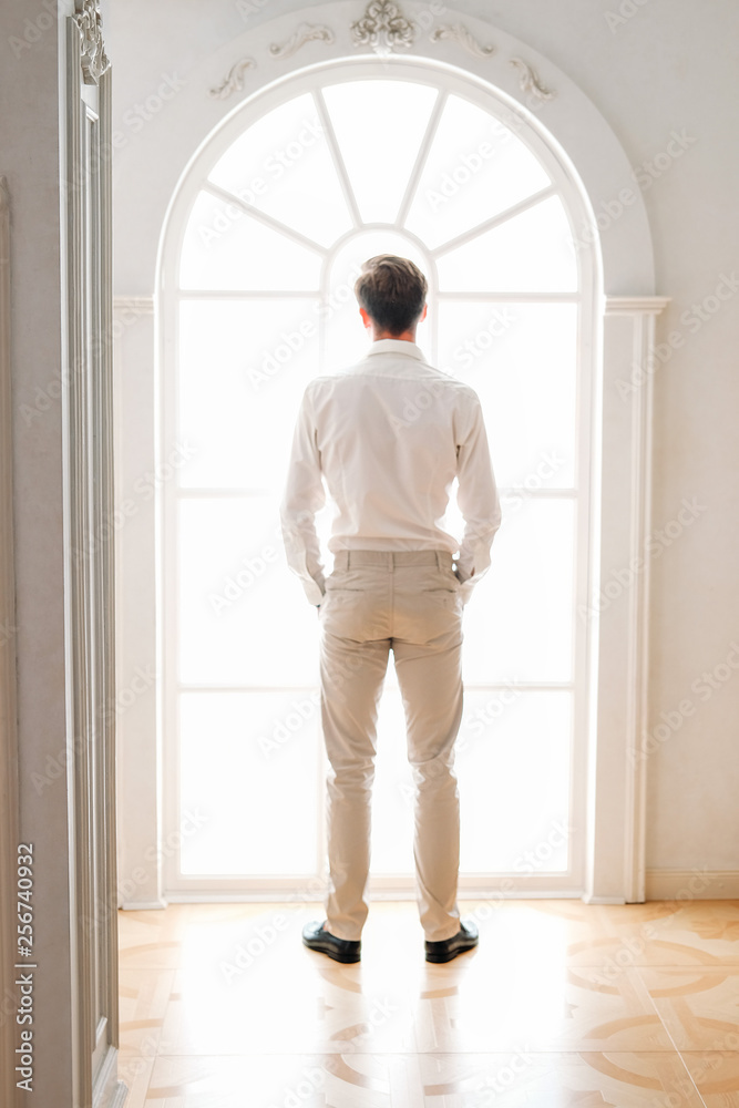 Young man standing face to window wearing light beige outfit Young man standing face to window wearing light beige outfit