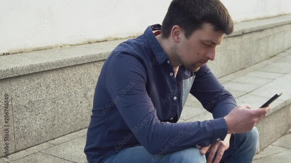 theme is man and social networks. young Caucasian sad male brunette in shirt is sitting in park in Letnec waiting, being late is using hand to hold mobile phone. Emotion resentment and disorder theme is man and social networks. young Caucasian sad male brunette in shirt is sitting in park in Letnec waiting, being late is using hand to hold mobile phone. Emotion resentment and disorder