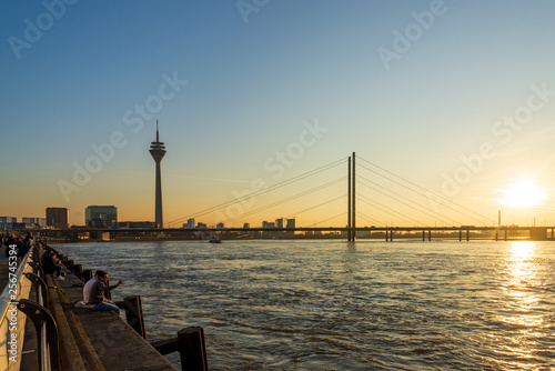 Outdoor scenery of people walk and sit for enjoy nice atmosphere at promenade along riverside of Rhine River during evening sunset time in Düsseldorf, Germany. © Peeradontax