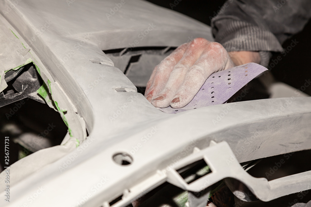 Obraz premium A man prepares a car body element for painting after an accident with the help of abrasive paper in a car repair shop. Recovery bumper after a collision.