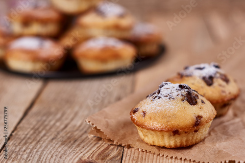 The two cupcakes with chocolate chips with powdered sugar lie on a kraft paper next to other cupcakes in a black baking sheet, on a wooden background. Close-up