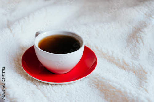 Big white morning cup of coffee and red plate in bed with pillow and cosy blanket.