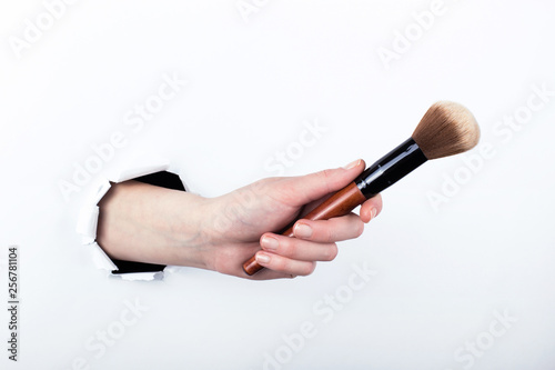 Female hand out of a hole in paper, holding a large powder brush. Isolate on white background.