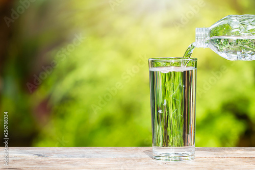 Pouring drinking water from bottle into glass on wooden tabletop on blurred fresh green nature background