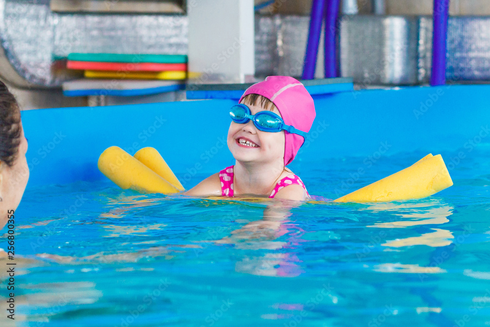 A little girl in a pink rubber hat and blue glasses swims in the pool on a yellow arc and laughs.