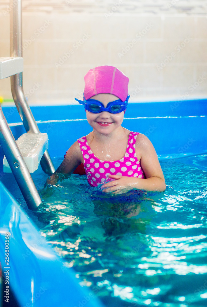 A little girl in a pink rubber hat and blue glasses in the pool at the stairs laughs.