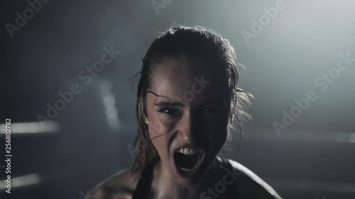 Portrait of tired female boxer standing on the boxing ring, looking intensely at the camera and screaming photo