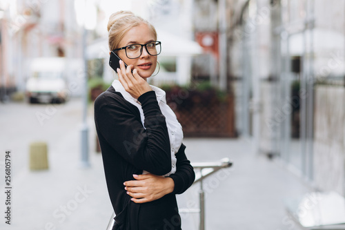 Beautiful trendy blond business woman in glasses talking by mobile phone and standing outside a office building.