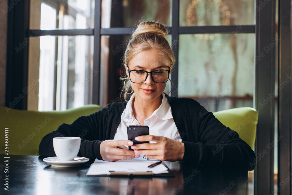 Blond business woman sitting on workspace and using laptop at office, she looks at a folder with documents, talking by phone and drinking cup of coffee. Blond business woman sitting on workspace and using laptop at office, she looks at a folder with documents, talking by phone and drinking cup of coffee.