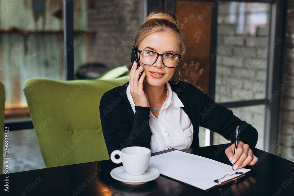 Blond business woman sitting on workspace and using laptop at office, she looks at a folder with documents, talking by phone and drinking cup of coffee. Blond business woman sitting on workspace and using laptop at office, she looks at a folder with documents, talking by phone and drinking cup of coffee.