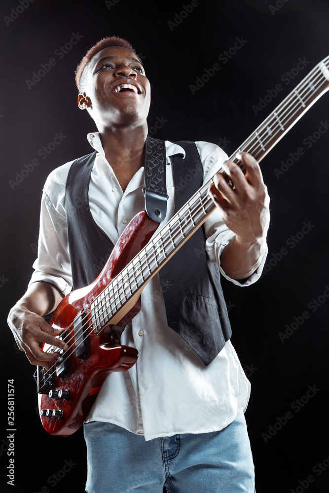 Obraz premium African American handsome jazz musician playing bass guitar in the studio on a black background. Music concept. Young joyful attractive guy improvising. Close-up retro portrait.