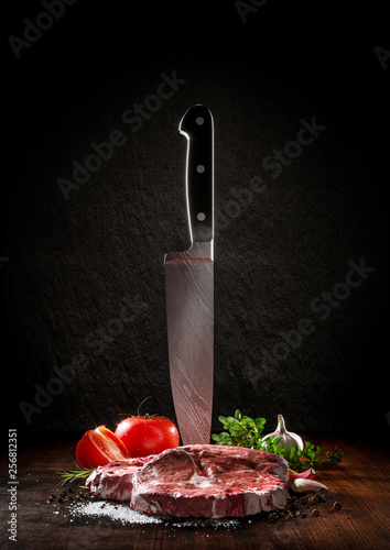 Beef rare steak on a wood desk with chef knife. Around are fresh tomatoes, sweet and spicy peppers, greens and spices, white salt and black pepper. 
