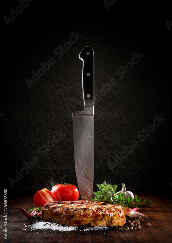 Beef steak on a wood desk with chef knife. Around are fresh tomatoes, sweet and spicy peppers, greens and spices, white salt and black pepper. Creative composition