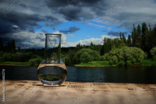 Clean water in a glass laboratory flask on wooden table on mountain river background. Ecological concept, the protection of water resources, the test of purity and quality of water.