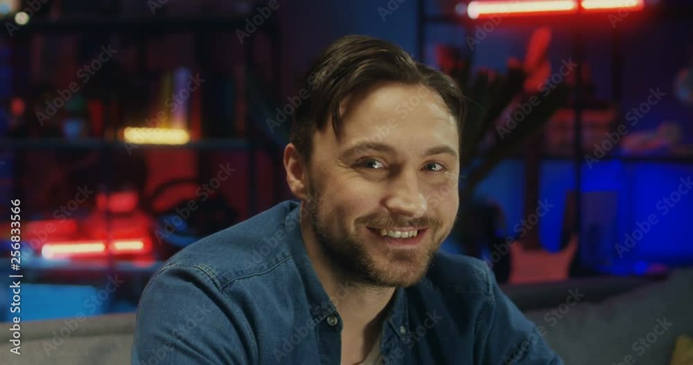 Close up of the Caucasian attractive young man turning his head to the camera and smiling cheerfully in the dark room at night. Portrait. Close up of the Caucasian attractive young man turning his head to the camera and smiling cheerfully in the dark room at night. Portrait.