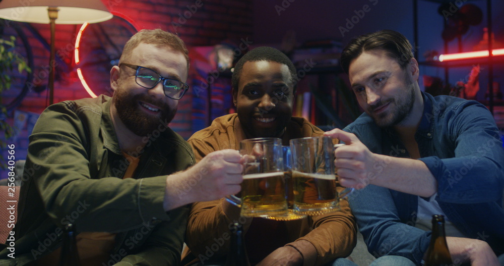 Portrait of the three multiethnic young attractive men smiling to the camera cheerfully with beer in hands in the cozy living room in the evening.