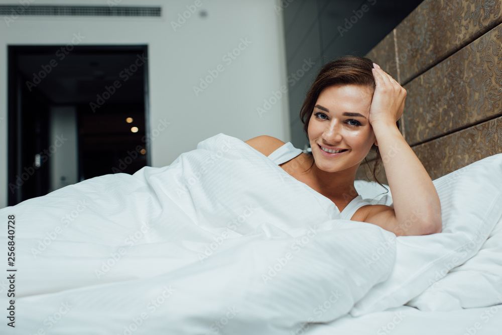 cheerful brunette woman smiling while lying on pillows under blanket in bed cheerful brunette woman smiling while lying on pillows under blanket in bed