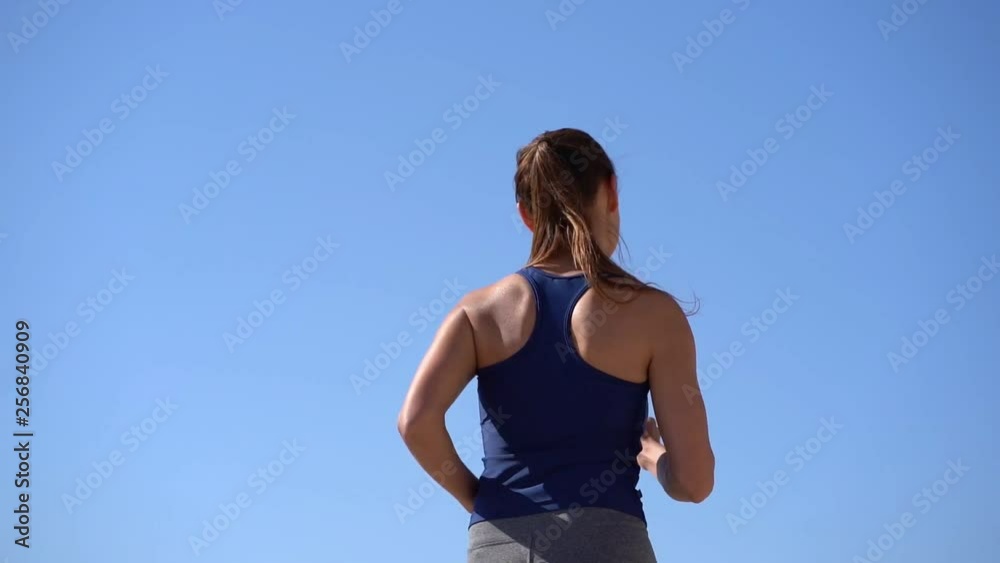 Back view of sporty girl jogging at sunny day. Low angle view of young sportswoman running against blue sky, slow motion. Workout concept Back view of sporty girl jogging at sunny day. Low angle view of young sportswoman running against blue sky, slow motion. Workout concept