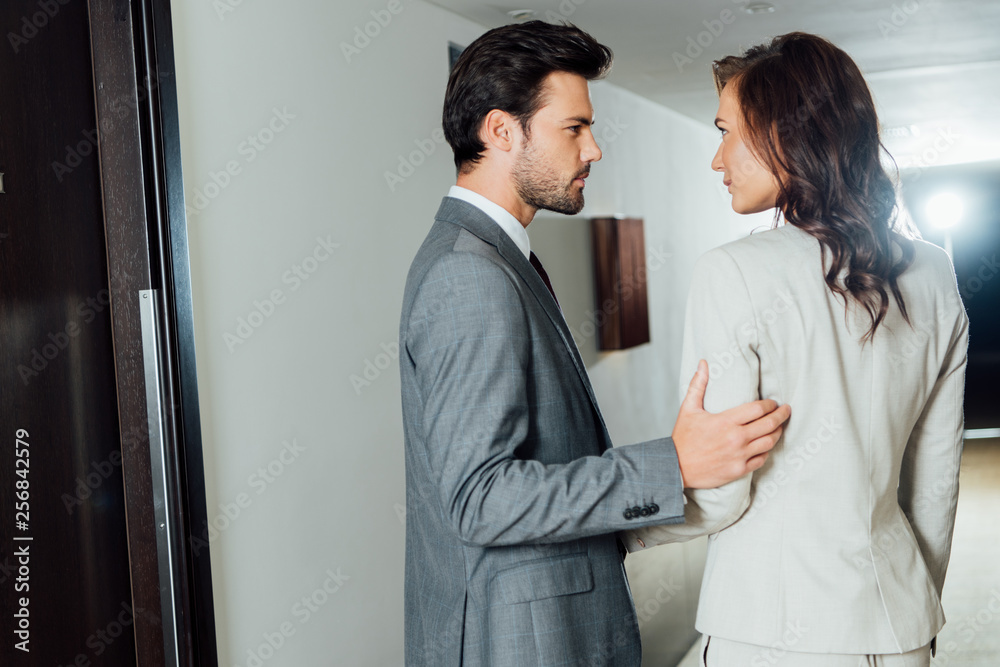 confident businessman touching hand of attractive businesswoman in formal wear while standing in hotel corridor