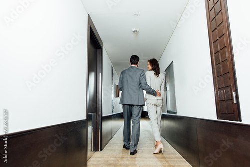 back view of confident businessman hugging businesswoman in formal wear while walking in hotel corridor