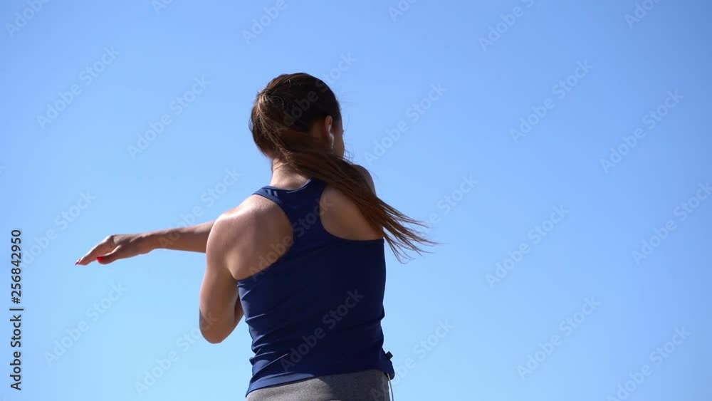 Back view of sporty girl stretching arms outdoor. Rear view of athletic young woman in sportswear and earphones exercising against blue sky at sunny day, low angle view. Workout concept Back view of sporty girl stretching arms outdoor. Rear view of athletic young woman in sportswear and earphones exercising against blue sky at sunny day, low angle view. Workout concept