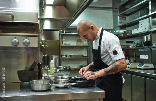 Food decoration. Side view of confident male chef with several tattoos on his arms garnishing his dish in a restaurant kitchen