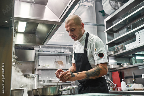 Important moment. Serious professional chef in apron, with several tattoos on his hands cooking in a restaurant kitchen