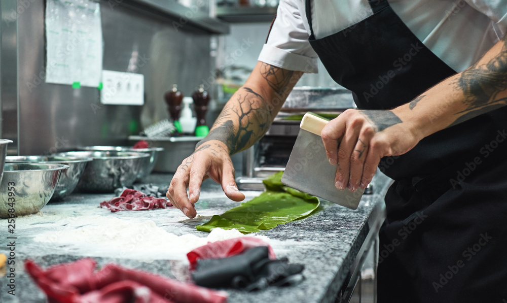 Secret recipe. Cropped photo of chef's hands with tattoos cutting dough on the kitchen table with flour for homemade italian pasta