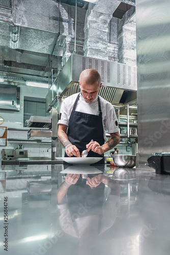 Working in a restaurant. Vertical portrait of professional male chef with tattoos on his arms garnishing his dish on the white plate