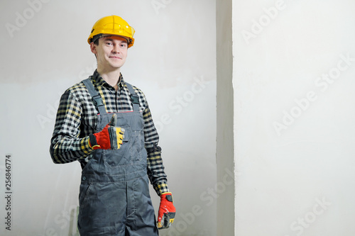 Portrait of a young male builder and repairman in a yellow helmet against the background of a wall.
