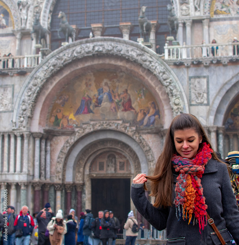 Venice / Italy 19 february 2019 : portrait of woman,she has long brown hair and she is wearing a grey coat and a multicolor scarf she is in front of the church and looks to the ground, people walking © Giorgio Tzitzi