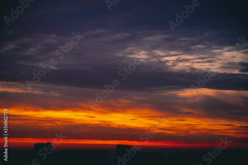 Cityscape with wonderful varicolored fiery dawn. Amazing dramatic multicolored cloudy sky. Dark silhouettes of city building roofs. Atmospheric background of sunrise in overcast weather. Copy space.