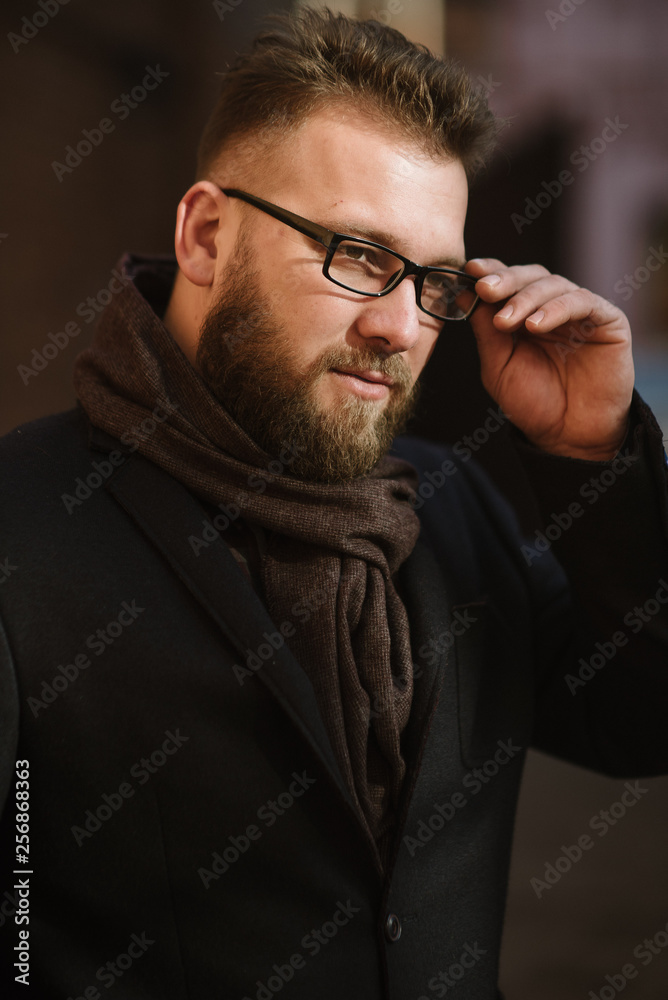 Fototapeta premium A man with a beard in a black warm coat with a scarf poses to advertise men's clothing in the winter. Advertise men's clothing