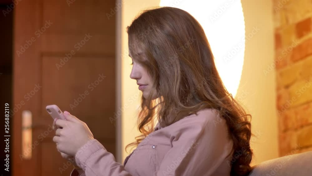 Portrait in profile of young teenage girl watching attentively into smartphone and adjusting her hair in cozy home atmosphere. Portrait in profile of young teenage girl watching attentively into smartphone and adjusting her hair in cozy home atmosphere.